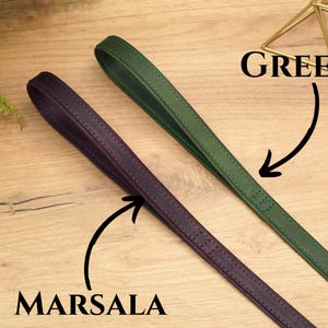 Puede incluir: Dos correas de cuero, una verde y otra morado oscuro, etiquetadas como "GREEN" y "MARSALA" respectivamente. Las correas est&aacute;n sobre una superficie de madera.