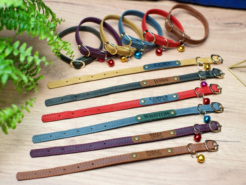 K&ouml;nnte beinhalten: Neun bunte Lederkatzenhalsb&auml;nder mit personalisierten Namensschildern und einem Gl&ouml;ckchen-Anh&auml;nger. Die Halsb&auml;nder sind in verschiedenen Farben, darunter Braun, Rot, Blau, Gr&uuml;n und Lila. Die Namensschilder sind mit den Namen "Cory", "Jessie", "Copper", "Winston", "Kelly", "Polly" und anderen Namen graviert.