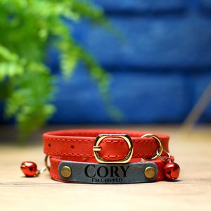 K&ouml;nnte beinhalten: Rotes Lederhalsband f&uuml;r Katzen mit einem grauen Lederanh&auml;nger, auf dem "Cory I'm chipped" steht. Das Halsband hat zwei goldene Gl&ouml;ckchen.