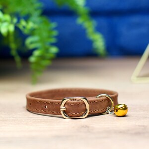 K&ouml;nnte beinhalten: Ein braunes Lederhalsband f&uuml;r Katzen mit einer goldenen Metallschnalle und einem goldenen Gl&ouml;ckchen-Anh&auml;nger.