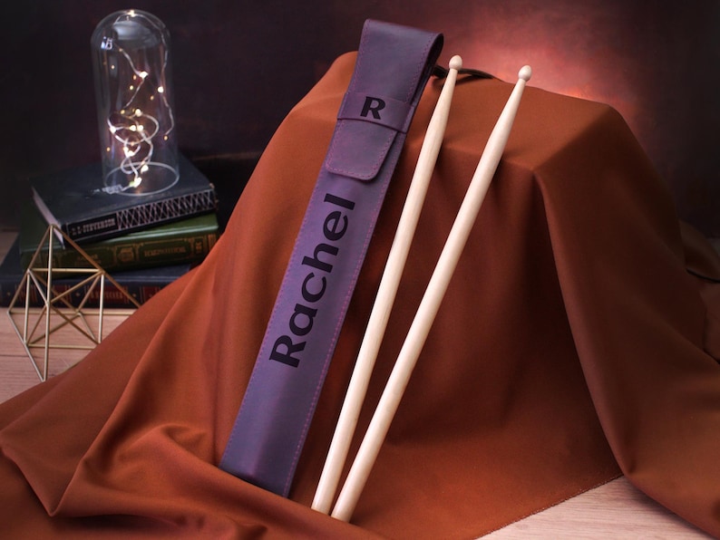 Puede incluir: Estuche de baquetas de cuero morado con el nombre "Rachel" grabado en &eacute;l. El estuche est&aacute; cerrado y dos baquetas de madera sobresalen de la parte superior.