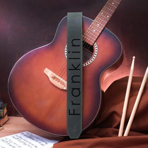 May include: A dark green leather guitar strap with the word "Franklin" printed in white on it. The strap is hanging on a brown acoustic guitar.