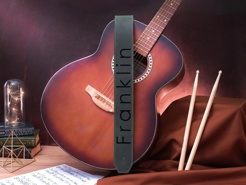 May include: A dark green leather guitar strap with the word "Franklin" printed in white on it. The strap is hanging on a brown acoustic guitar.