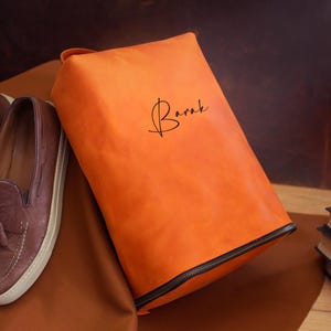 May include: An orange leather shoe bag with a black zipper and the name "Barak" in cursive script. Beside the bag are a pair of brown leather loafers with tassels and a stack of books.