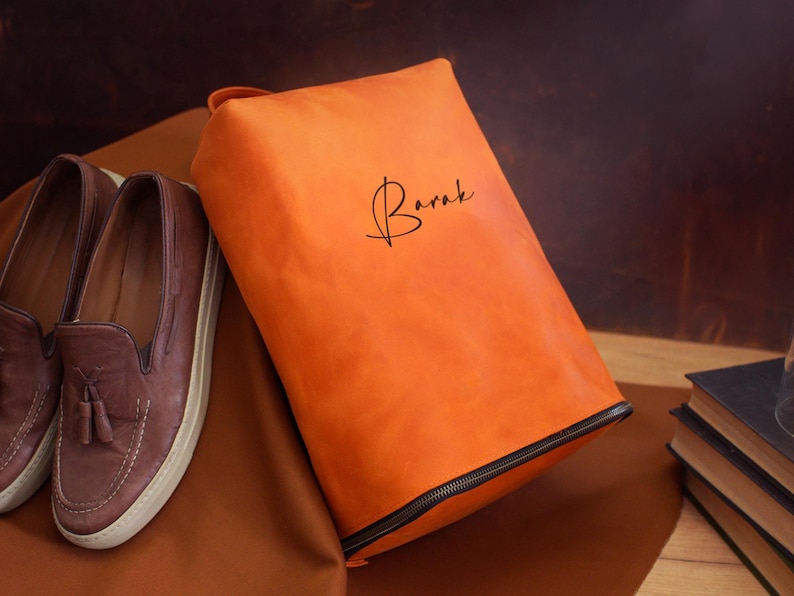 May include: An orange leather shoe bag with a black zipper and the name "Barak" in cursive script. Beside the bag are a pair of brown leather loafers with tassels and a stack of books.