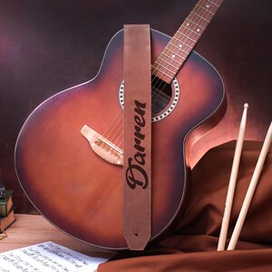 May include: A brown leather guitar strap with the name "Darren" engraved on it. The strap is attached to an acoustic guitar with a brown wood body and a natural wood fretboard.
