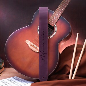 May include: A purple leather guitar strap with the name "Maximilian" written in white script. The strap is attached to an acoustic guitar with a brown wood body and a natural wood fretboard.