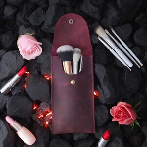 May include: A burgundy leather makeup brush holder with a snap closure. The holder is filled with makeup brushes and is sitting on a bed of black rocks. There are two pink roses and two red lipsticks on the rocks.
