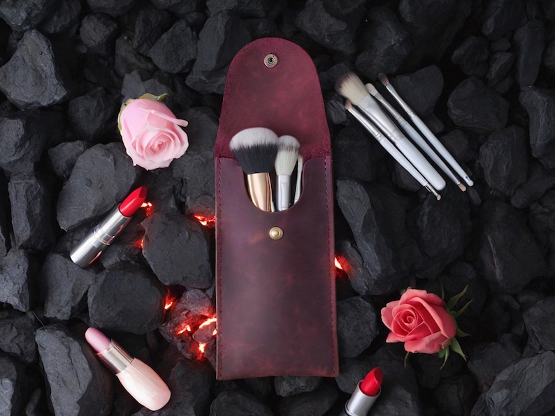 May include: A burgundy leather makeup brush holder with a snap closure. The holder is filled with makeup brushes and is sitting on a bed of black rocks. There are two pink roses and two red lipsticks on the rocks.
