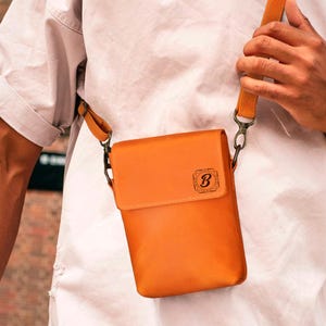 May include: A rectangular, orange crossbody bag with a flap closure and a long, adjustable strap. The bag features a small, embossed "B" logo on the front. The bag is made of a smooth material and has metal hardware.