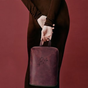 May include: A burgundy-colored, rectangular handbag with a top handle. The bag has the name "Bela" in a cursive script on the front. The background is a solid, deep red color.