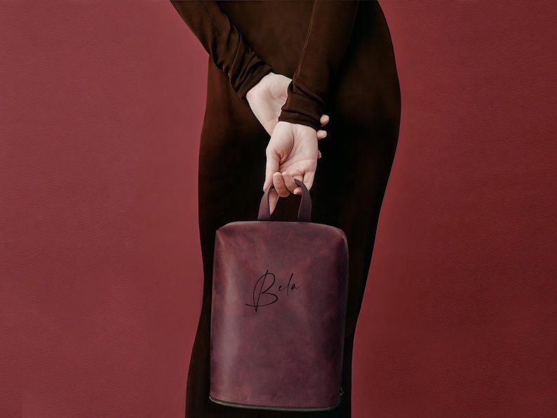 May include: A burgundy-colored, rectangular handbag with a top handle. The bag has the name "Bela" in a cursive script on the front. The background is a solid, deep red color.