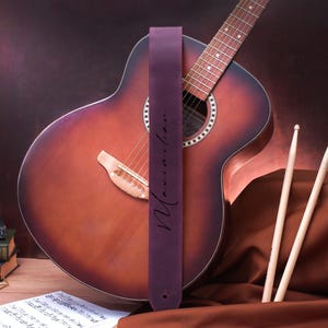 May include: A purple leather guitar strap with the name "Maximilian" written in white script. The strap is attached to an acoustic guitar with a brown wood body and a natural wood fretboard.