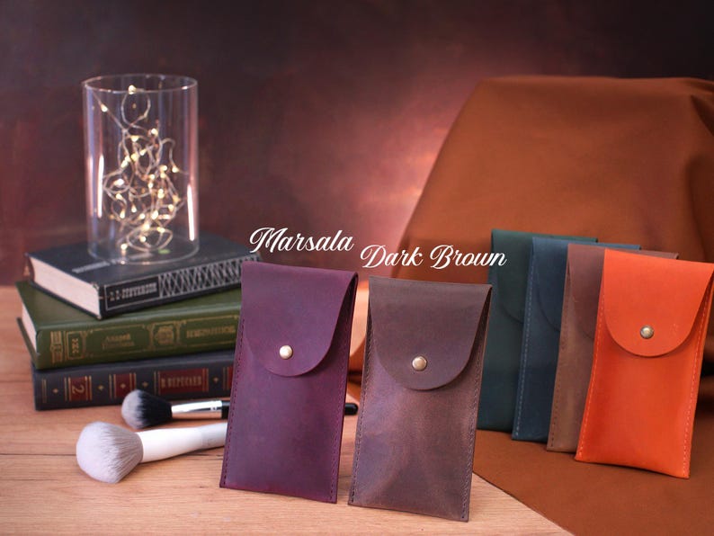 May include: Four leather pouches in different colors, including marsala, dark brown, navy blue, and orange. The pouches have a flap with a button closure. The text "Marsala Dark Brown" is written on the image.