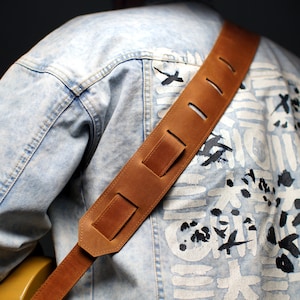 May include: A brown leather guitar strap with adjustable length and multiple holes. The strap is worn over a light blue denim jacket with a white and black abstract design.