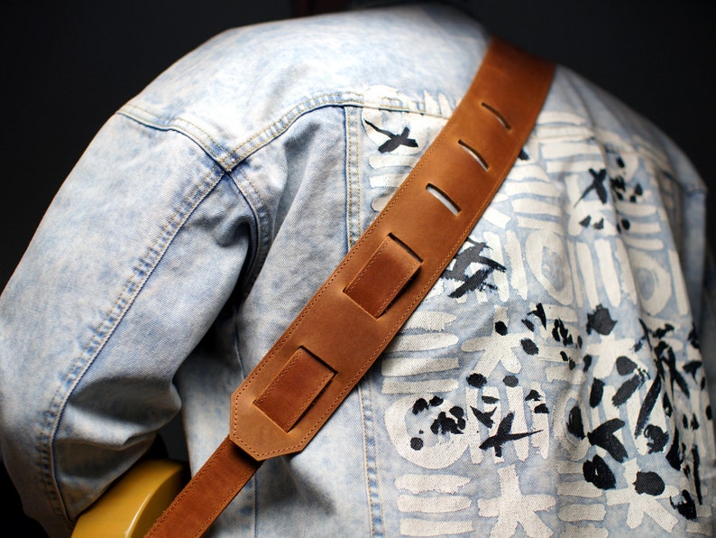 May include: A brown leather guitar strap with adjustable length and multiple holes. The strap is worn over a light blue denim jacket with a white and black abstract design.