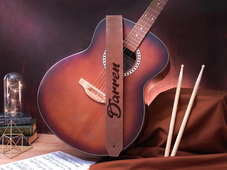 May include: A brown leather guitar strap with the name "Darren" engraved on it. The strap is attached to an acoustic guitar with a brown wood body and a natural wood fretboard.