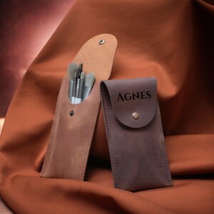 May include: Two leather makeup brush holders. One is brown and has a set of brushes inside. The other is dark brown and has the name Agnes engraved on the front.