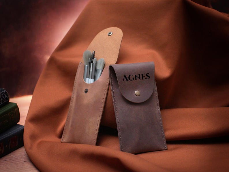 May include: Two leather makeup brush holders. One is brown and has a set of brushes inside. The other is dark brown and has the name Agnes engraved on the front.