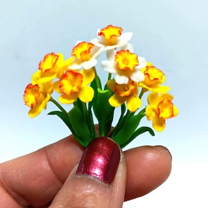 Puede incluir: Un ramo de narcisos amarillos y blancos con bordes rojos en los pétalos. Las flores están hechas de arcilla polimérica y están dispuestas en un racimo con hojas verdes.