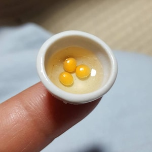Könnte beinhalten: Eine kleine weiße Keramikschüssel mit drei gelben Eigelb darin. Die Schüssel liegt auf einem Finger.