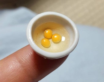 May include: A small white ceramic bowl with three yellow egg yolks inside. The bowl is sitting on a finger.