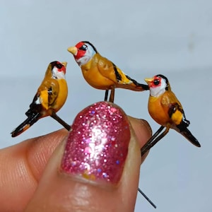 May include: Three miniature goldfinch figurines, each featuring a yellow body, black and white wing markings, and a distinct red face patch. These small bird sculptures are attached to thin, dark metal stems, showcasing detailed painted features.