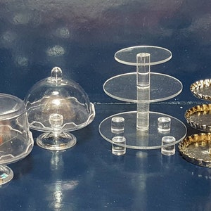 Könnte beinhalten: Drei Tortenständer, zwei aus klarem Kunststoff und einer aus goldenem Metall. Die klaren Ständer haben eine runde Basis und einen kuppelförmigen Deckel. Der goldene Ständer hat drei Etagen mit einem gewellten Rand.
