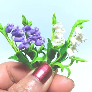 Könnte beinhalten: Ein Miniaturstrauß handgefertigter Blumen mit lila und weißen Blüten mit grünen Blättern und Stielen. Die Blumen werden in einer Hand mit rotem Nagellack gehalten.