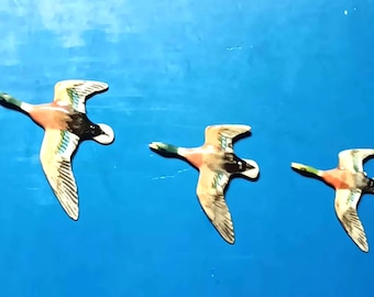May include: Three decorative duck figurines in flight against a bright blue background. The ducks feature detailed wings with a mix of colors, including green, brown, and white. The ducks are of varying sizes, creating a sense of depth.