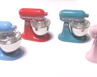 May include: Four miniature stand mixers in pastel colors: blue, red, teal, and pink. Each mixer has a silver mixing bowl and a matching colored base and top. The mixers are arranged on a white surface.
