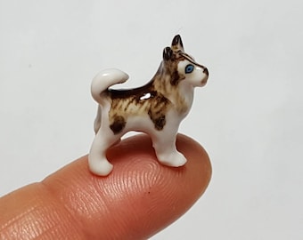 May include: A miniature ceramic Husky dog figurine. The small dog has a white body with brown and tan markings, a fluffy tail, and blue eyes. The figurine is standing on a finger, showcasing its small size.