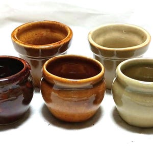 May include: Five small ceramic pots in various shades of brown and beige. The pots are all round and have a slightly flared lip.