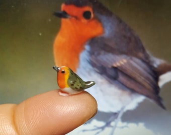 Op de afbeelding: Een miniatuur keramieken roodborstje met een rode borst, gele en groene vleugels en een witte buik. De kleine vogel zit op een vingertop, met een grotere, wazige afbeelding van een roodborstje op de achtergrond.