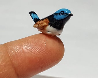 May include: A small ceramic figurine of a blue, black, and brown bird with a white belly. The bird is perched on a finger.