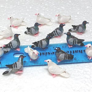 Może przedstawiać: Kolekcja miniaturowych figurek gołębi. Ptaki są w różnych pozach, niektóre w locie, a inne stoją. Gołębie są pomalowane w odcieniach szarości, czerni i bieli, z różowymi stopami i dziobami. Niebieska linijka znajduje się w tle.