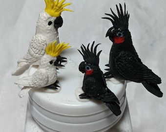 May include: Four small, hand-painted bird figurines. Two are white cockatoos with yellow crests, and two are black cockatoos with red accents. The figurines are arranged on a white surface.