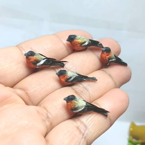 May include: A group of miniature bird ornaments. Each bird has a black head, a bright orange breast, and black and white wings. The birds are resting on a hand, highlighting their small size and fine details. These figurines are a delightful decorative item.