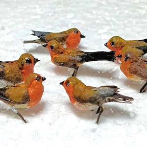 May include: A collection of seven miniature bird figurines. Each bird has a brown body, a red breast, and black and white tail feathers. The birds are standing on a white surface.