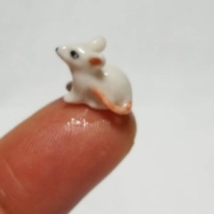 May include: A small white ceramic mouse figurine with pink ears and a tail. The figurine is sitting on a finger.