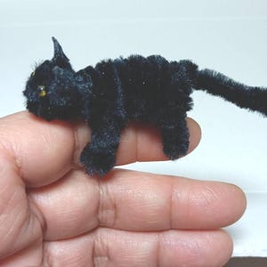 Könnte beinhalten: Eine kleine schwarze Katze aus Pfeifenreinigern. Die Katze ist auf einem Finger platziert, mit dem Schwanz um den Finger gewickelt.