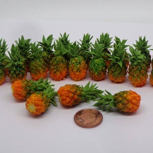 May include: A collection of 17 miniature pineapples with green tops and orange bodies. The pineapples are arranged in a semi-circle on a white surface.