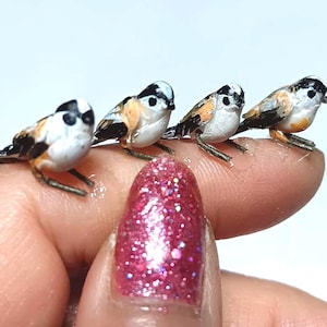Może przedstawiać: Cztery miniaturowe figurki ptaków z białymi, czarnymi i pomarańczowymi oznaczeniami siedzą na palcu. Ptaki mają szczegółowe cechy, w tym maleńkie nogi i dzioby. Palec ma różowy lakier do paznokci z brokatem.