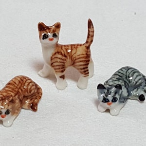 May include: Three ceramic cat figurines. One is brown and white with black stripes, standing on all four paws. Another is brown and white with black stripes, lying down. The third is grey and white with black stripes, lying down.