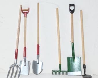 May include: A collection of miniature gardening tools, including a pitchfork, shovels, a rake, and a hoe. The tools have wooden handles with red and green accents and silver-colored metal blades. The tools are arranged against a white background.