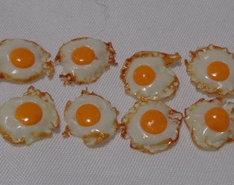 May include: Eight miniature fried eggs, each with a bright yellow yolk and a white, slightly browned egg white. The eggs are arranged in two rows of four on a white surface.