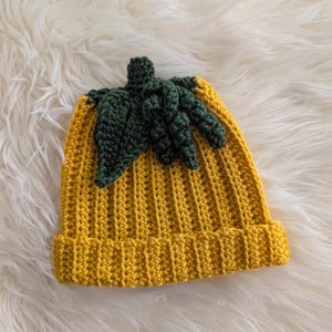 Puede incluir: Un gorro de ganchillo amarillo hecho a mano con un dise&ntilde;o de hoja y tallo verde. El gorro est&aacute; hecho de un hilo suave y tiene un borde acanalado. La hoja y el tallo verdes est&aacute;n unidos a la parte superior del gorro, creando una apariencia de calabaza.
