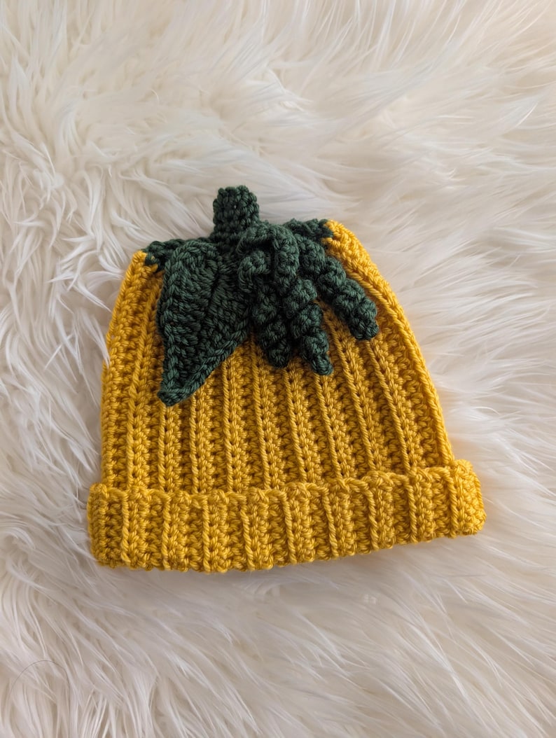 Puede incluir: Un gorro de ganchillo amarillo hecho a mano con un dise&ntilde;o de hoja y tallo verde. El gorro est&aacute; hecho de un hilo suave y tiene un borde acanalado. La hoja y el tallo verdes est&aacute;n unidos a la parte superior del gorro, creando una apariencia de calabaza.