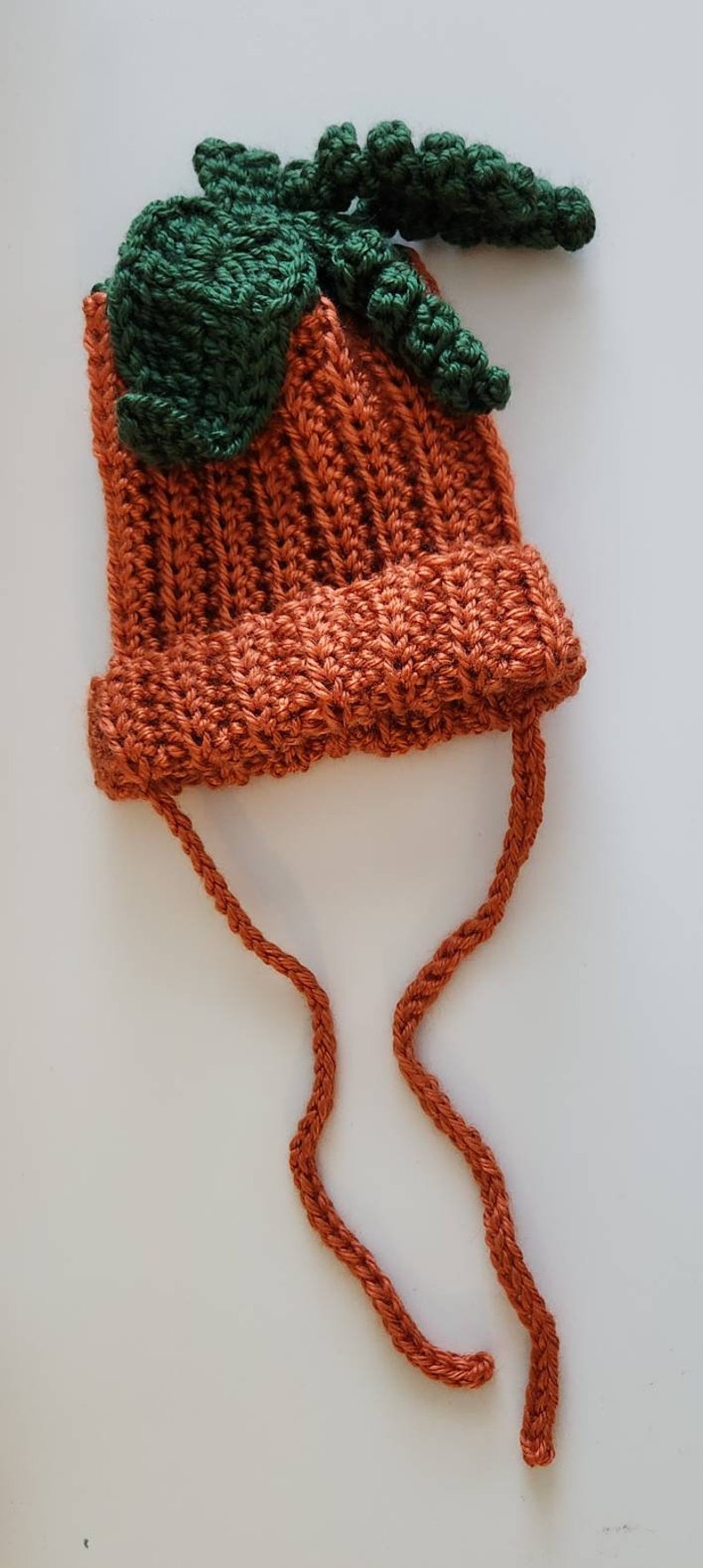 Puede incluir: Un gorro de calabaza de ganchillo hecho a mano en tonos naranja y verde. El gorro presenta un dise&ntilde;o acanalado y un detalle de hoja y tallo verde en la parte superior. Tiene dos lazos naranjas.