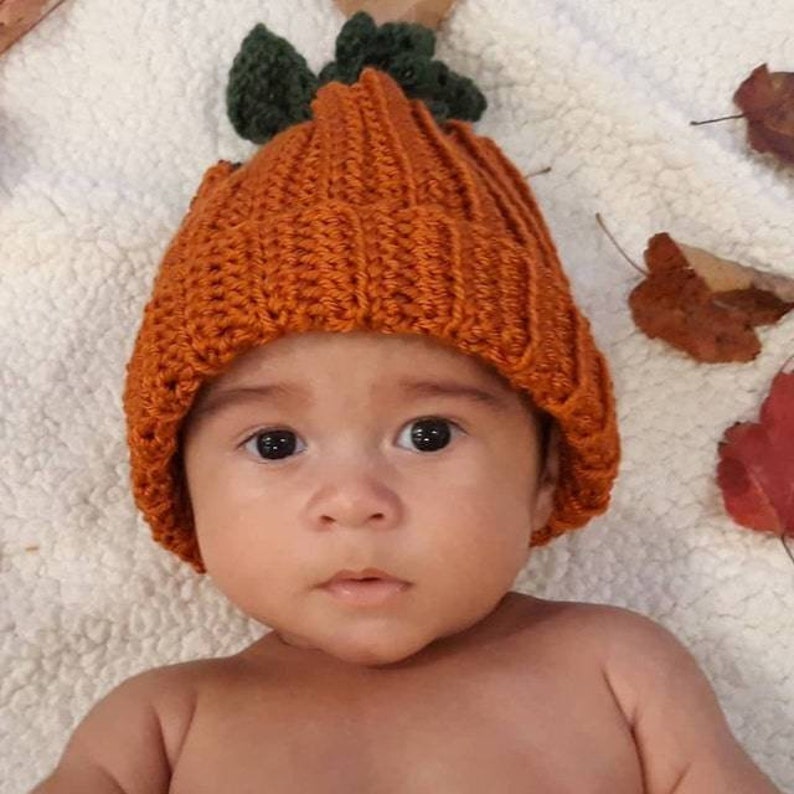 Puede incluir: Un beb&eacute; con un gorro de calabaza naranja tejido a ganchillo con hojas verdes. El gorro est&aacute; sobre una superficie blanca y texturizada con hojas de oto&ntilde;o.
